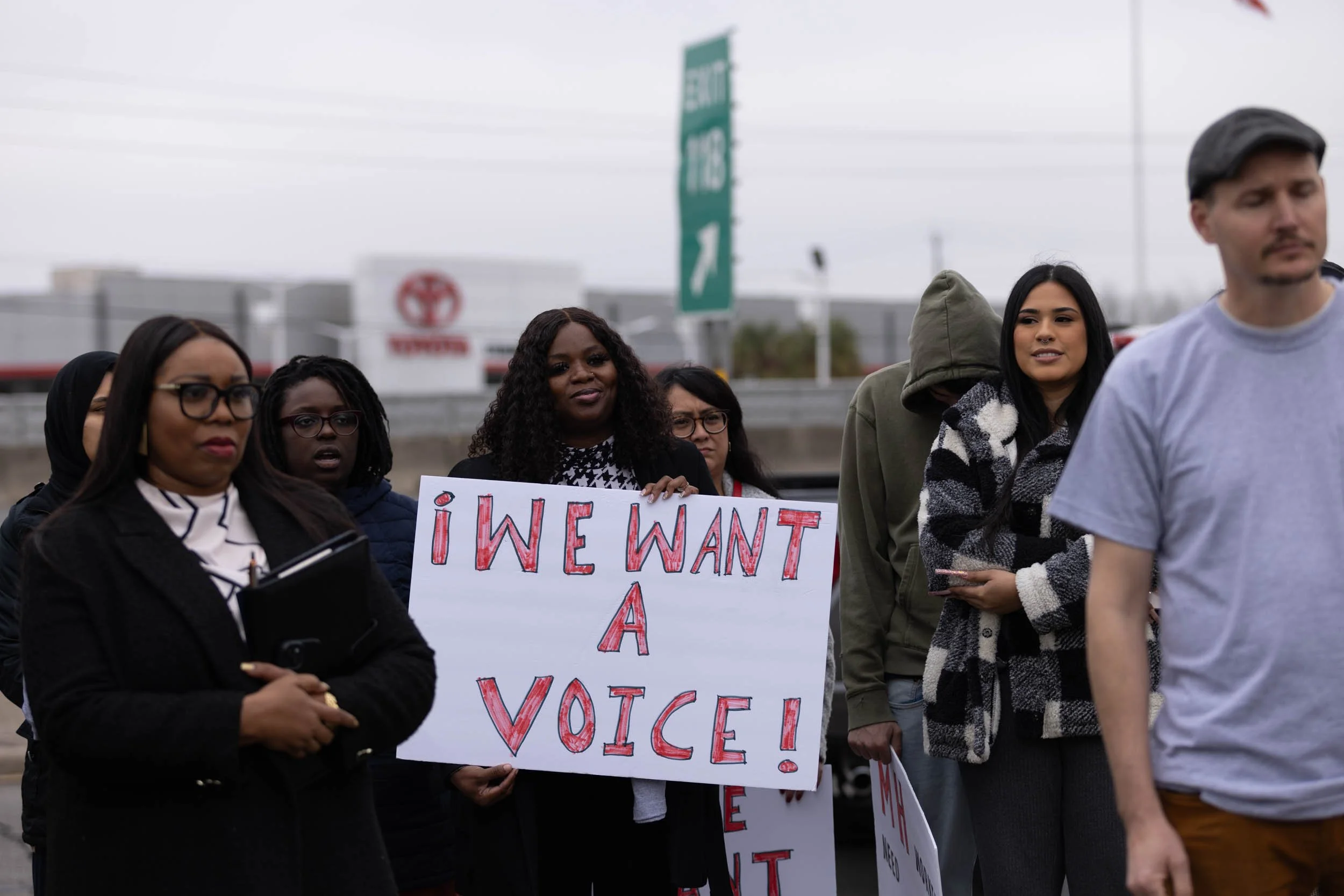 Image of a crowd at a rally for UWHC; in the center, a Black woman holds a white sign with red lettering that reads "¡We want a voice!"