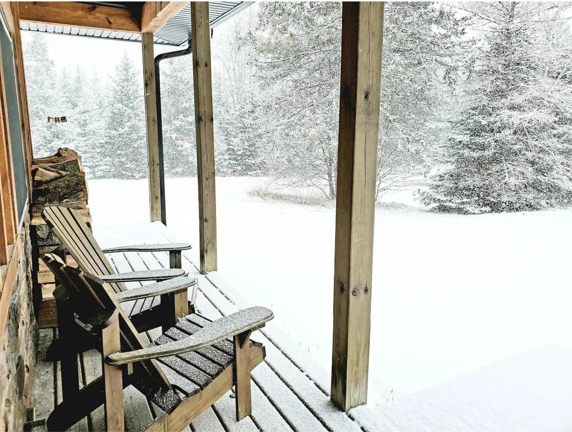 Image of a cottage porch with two chairs, with snow on the ground and trees.