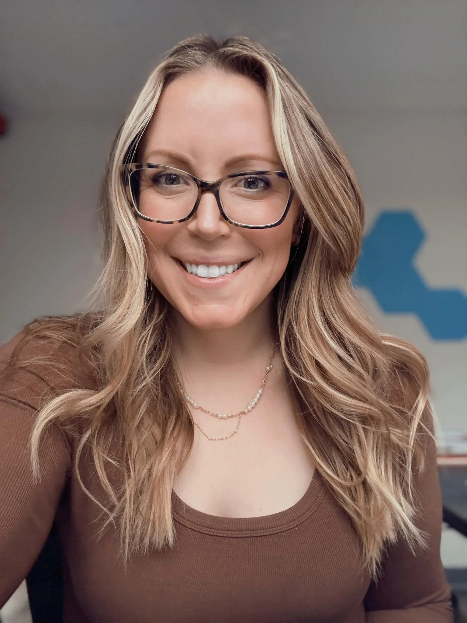 A woman with long blonde hair, wearing glasses and a brown top, smiling at the camera in an indoor setting.