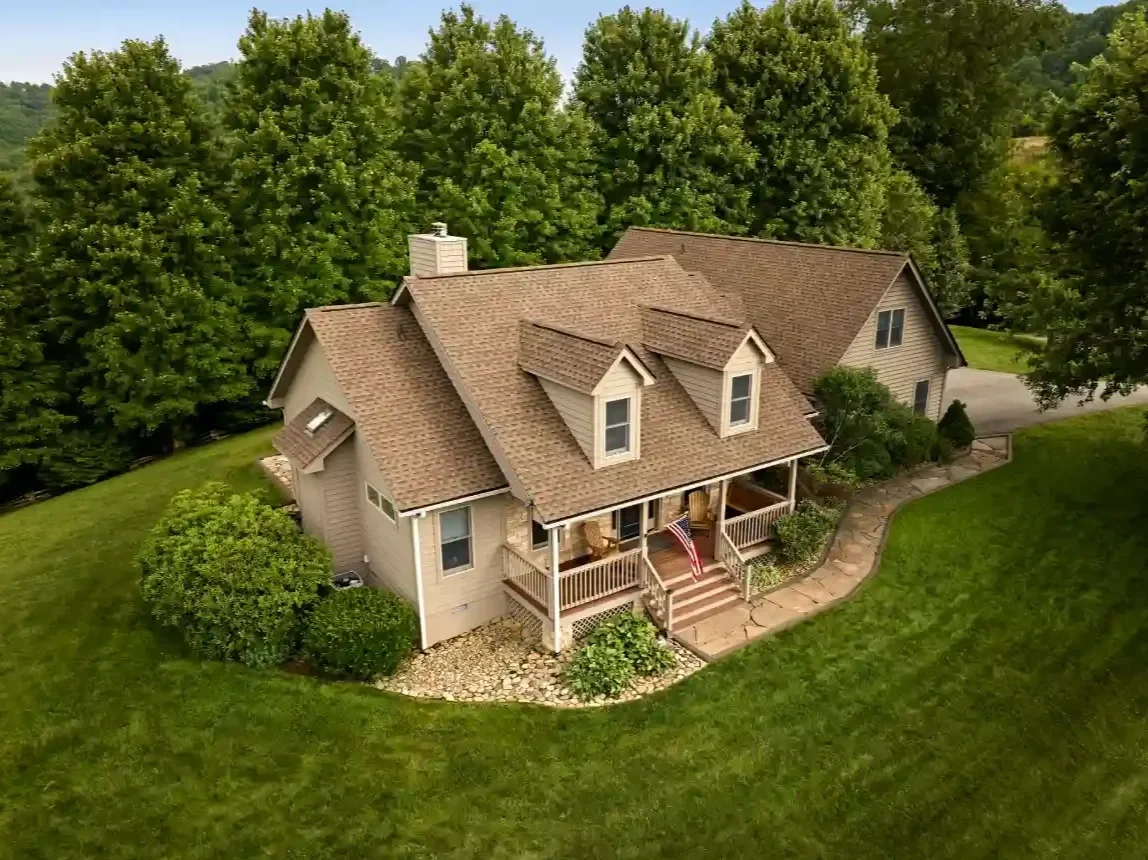 Aerial view of a two-story house with a brown shingle roof, beige siding, a front porch with chairs, and surrounded by a green lawn and trees.