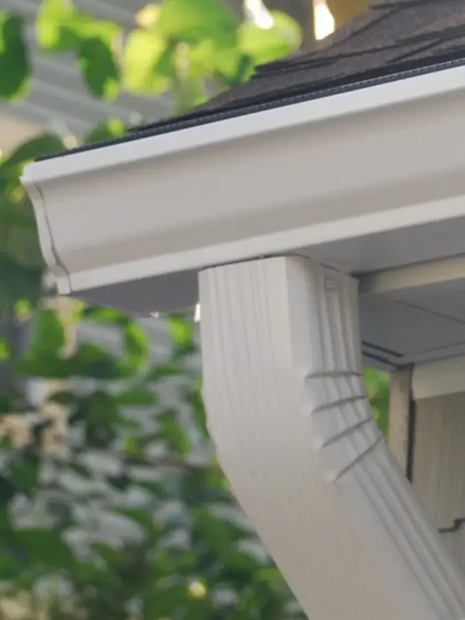 Close-up of a house's exterior corner showing white siding, gutter, and downspout, with green leaves in the background.