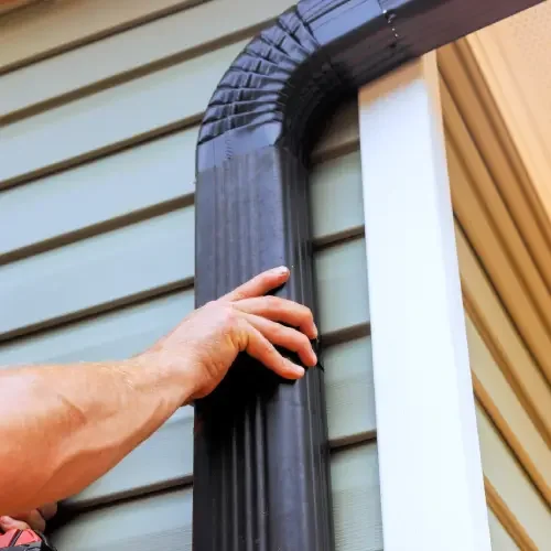 A person installing or adjusting a black rain gutter on the side of a house with beige siding.