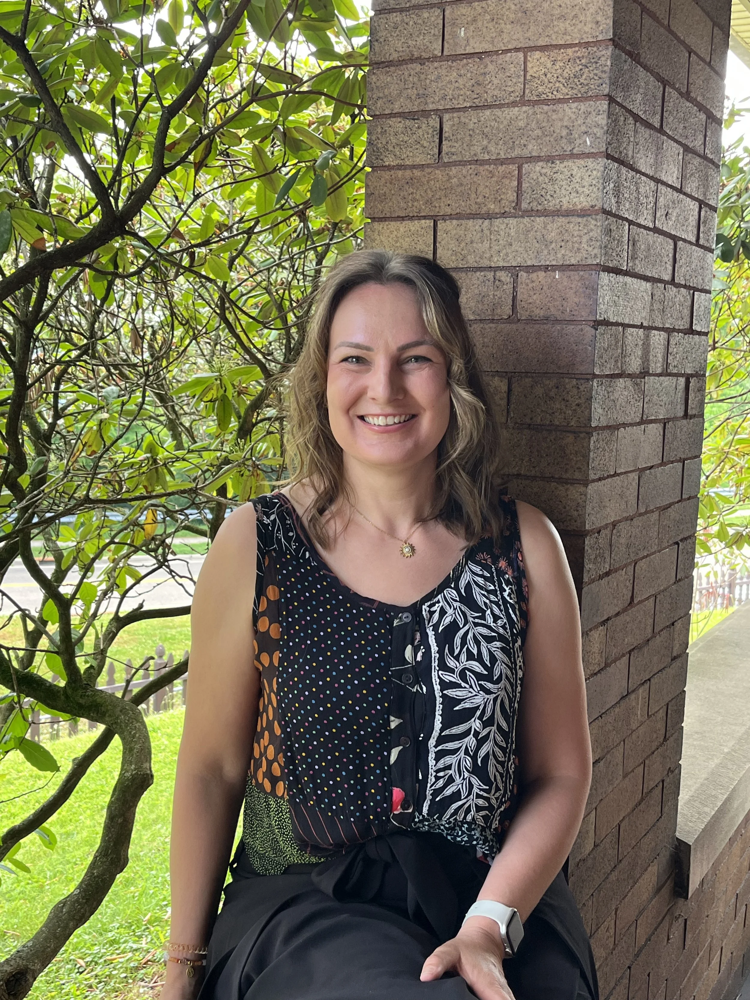 A woman with curly blonde hair wearing a black dress with colorful patterns, smiling and sitting outside near a brick wall and green foliage.