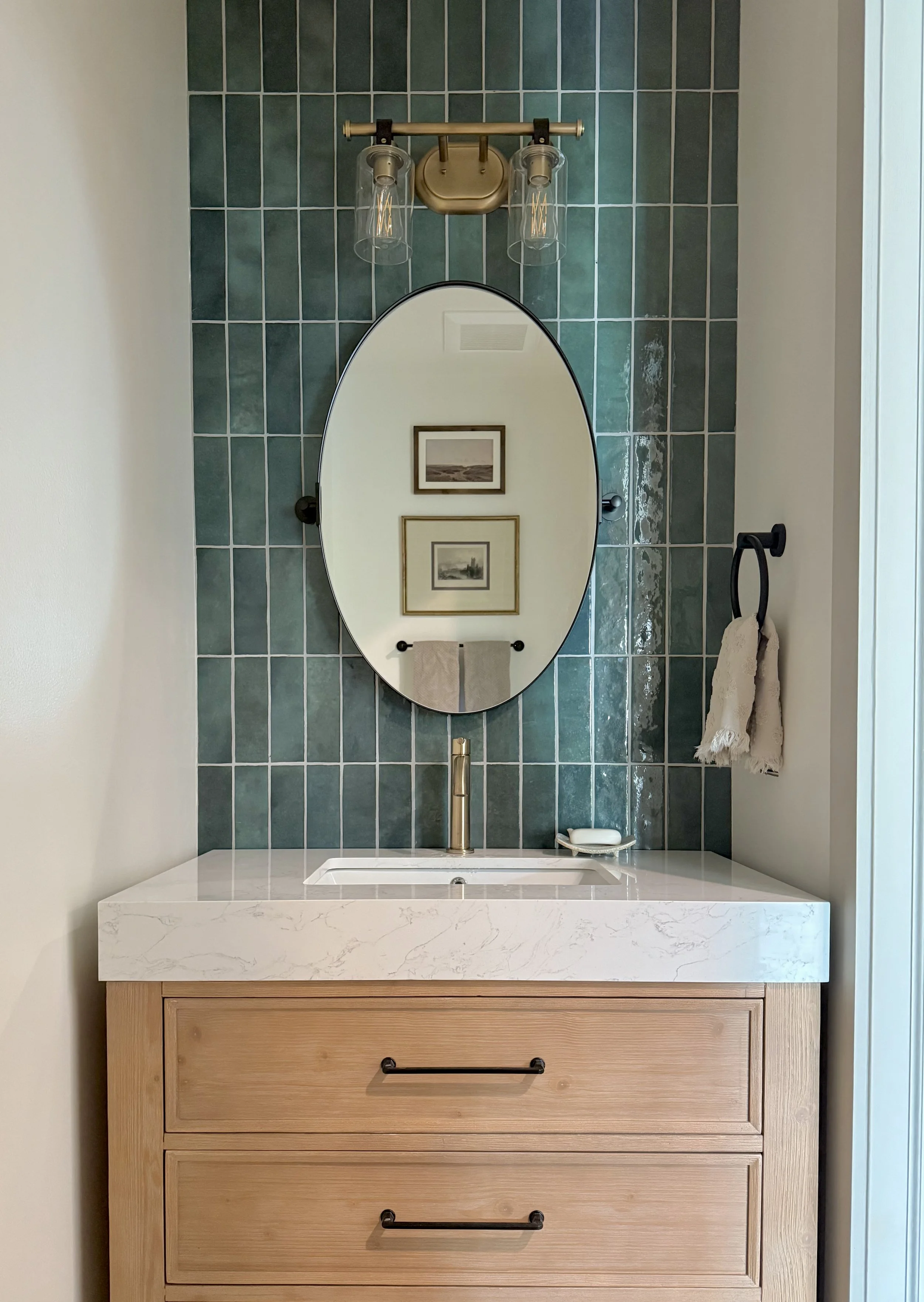 Bathroom vanity with a white marble top, wooden drawers, an oval mirror, a gold faucet, two framed pictures on the wall opposite, and a towel hanging from a black ring.