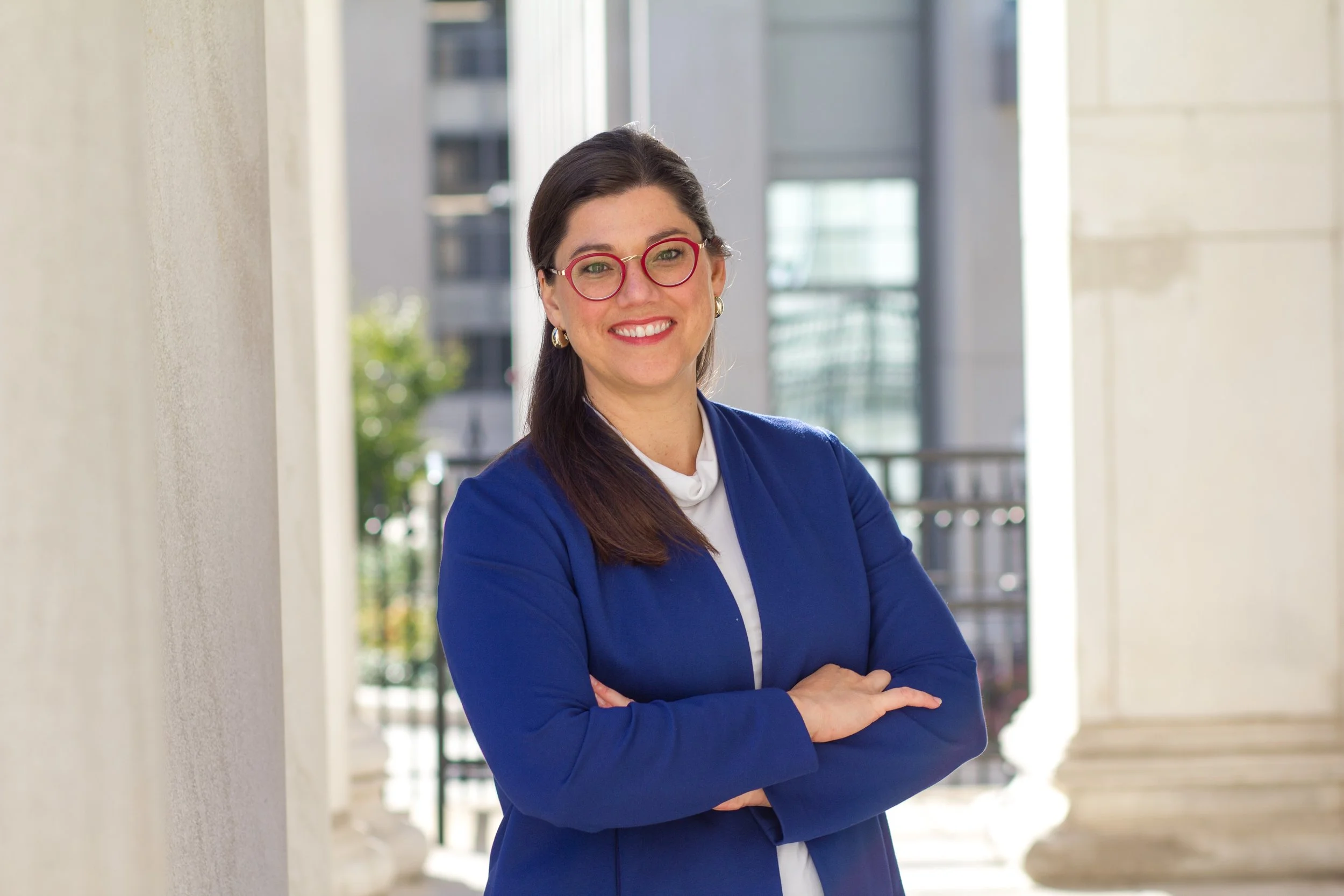 A woman wearing a blue blazer and red glasses smiling outdoors, standing with her arms crossed on a city balcony or walkway.