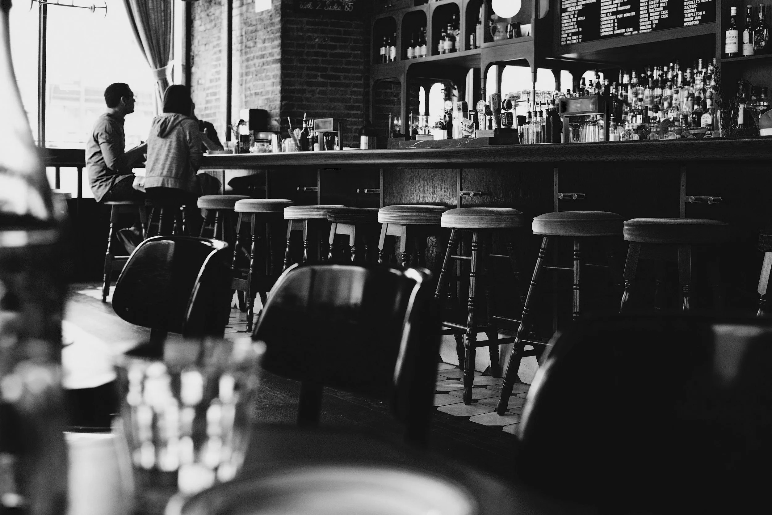 Black and white photo of a bar interior with bar stools, bottles on shelves, and two people sitting at the counter.