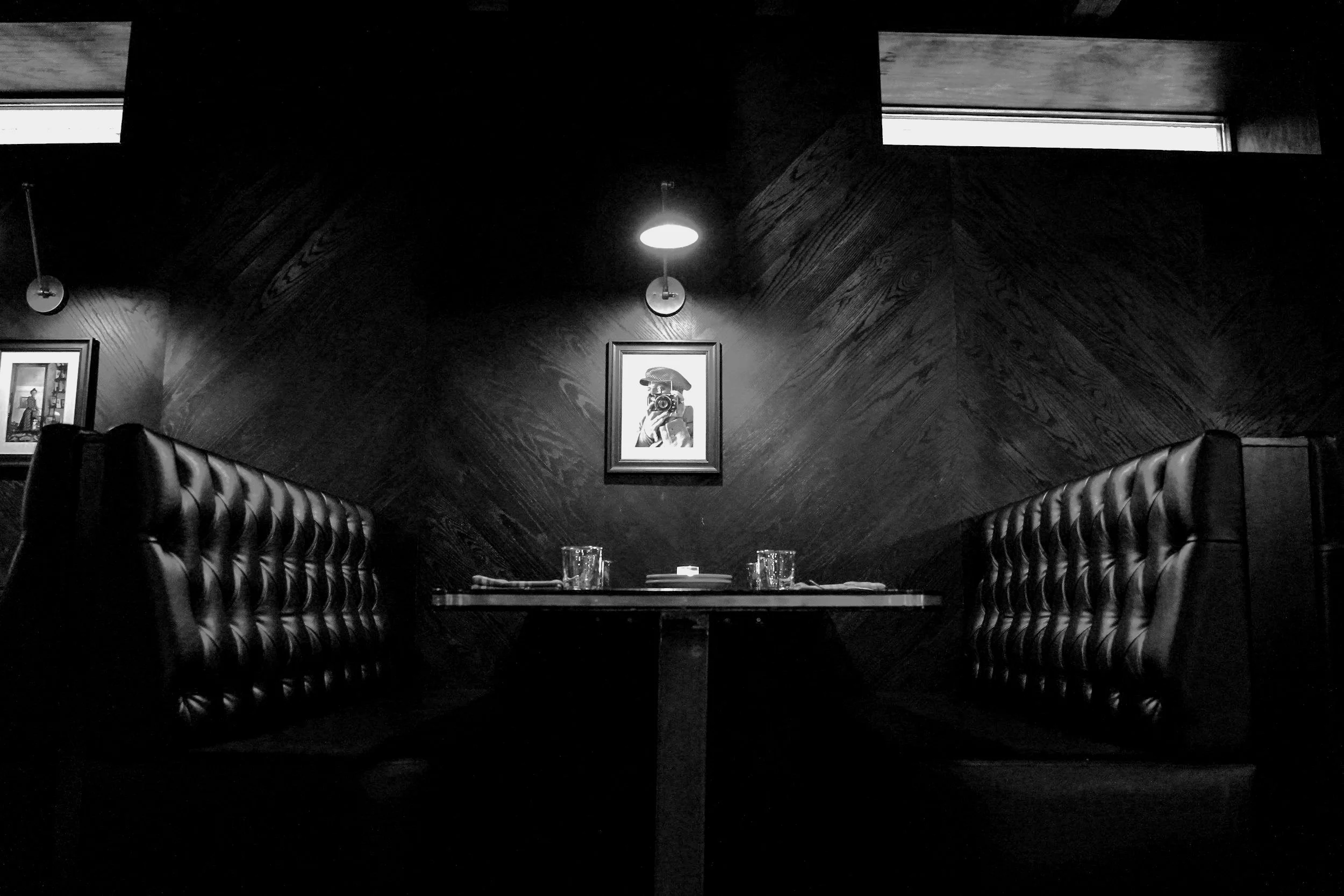 Black and white photo of an empty restaurant booth with tufted seats, featuring a central table set for two with glasses and napkins, and framed artwork on the dark patterned wall illuminated by wall lights.