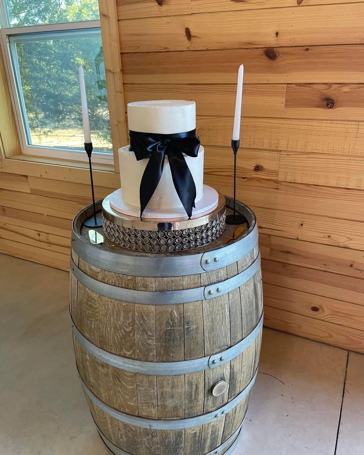 Two-tier wedding cake with black ribbon on wooden barrel with decorative stand and candles.