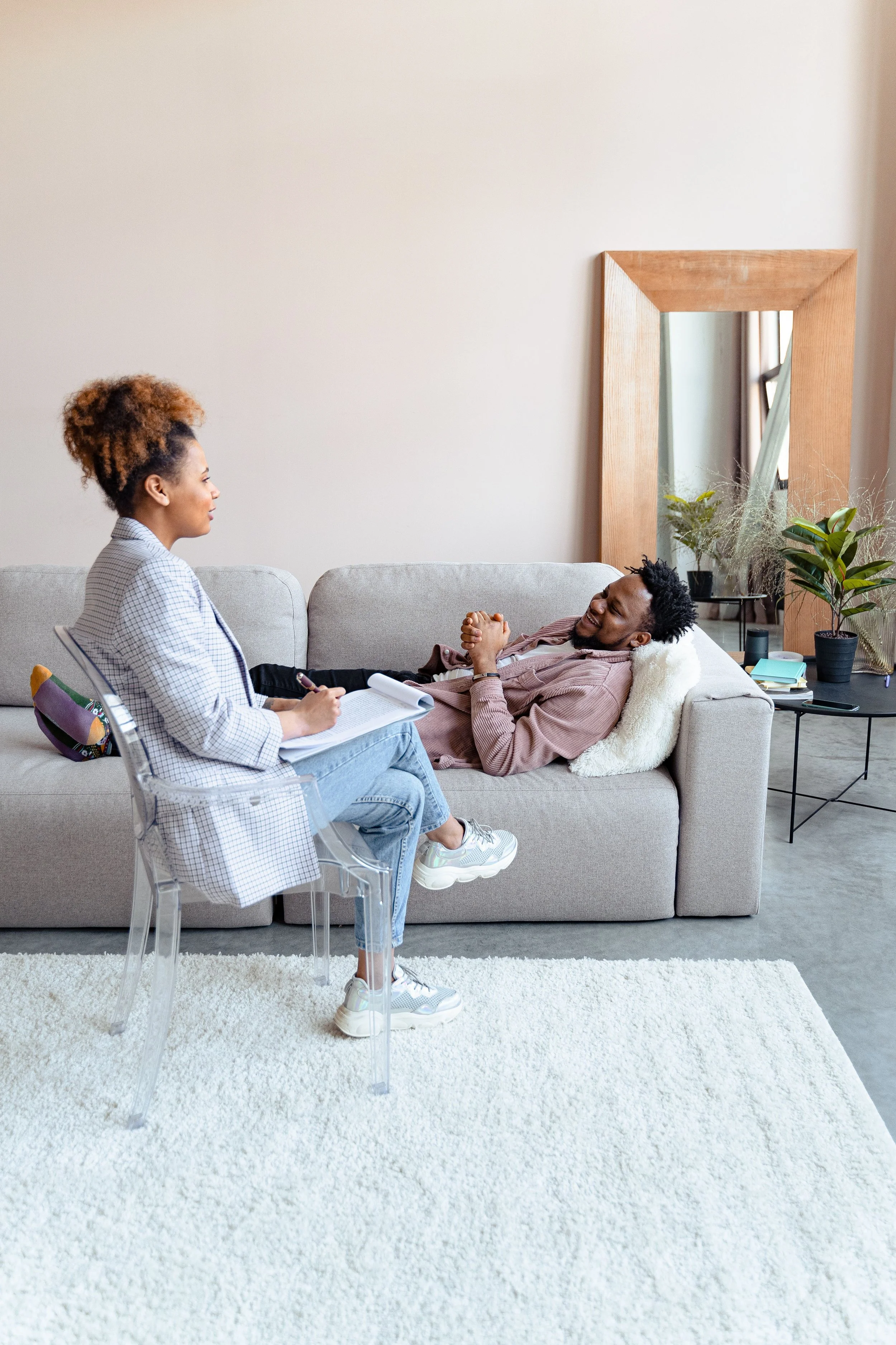 A woman and a man in a living room, seated on a sofa and talking. The woman is taking notes, and the man is smiling. There is a large mirror, among plants, in the background.