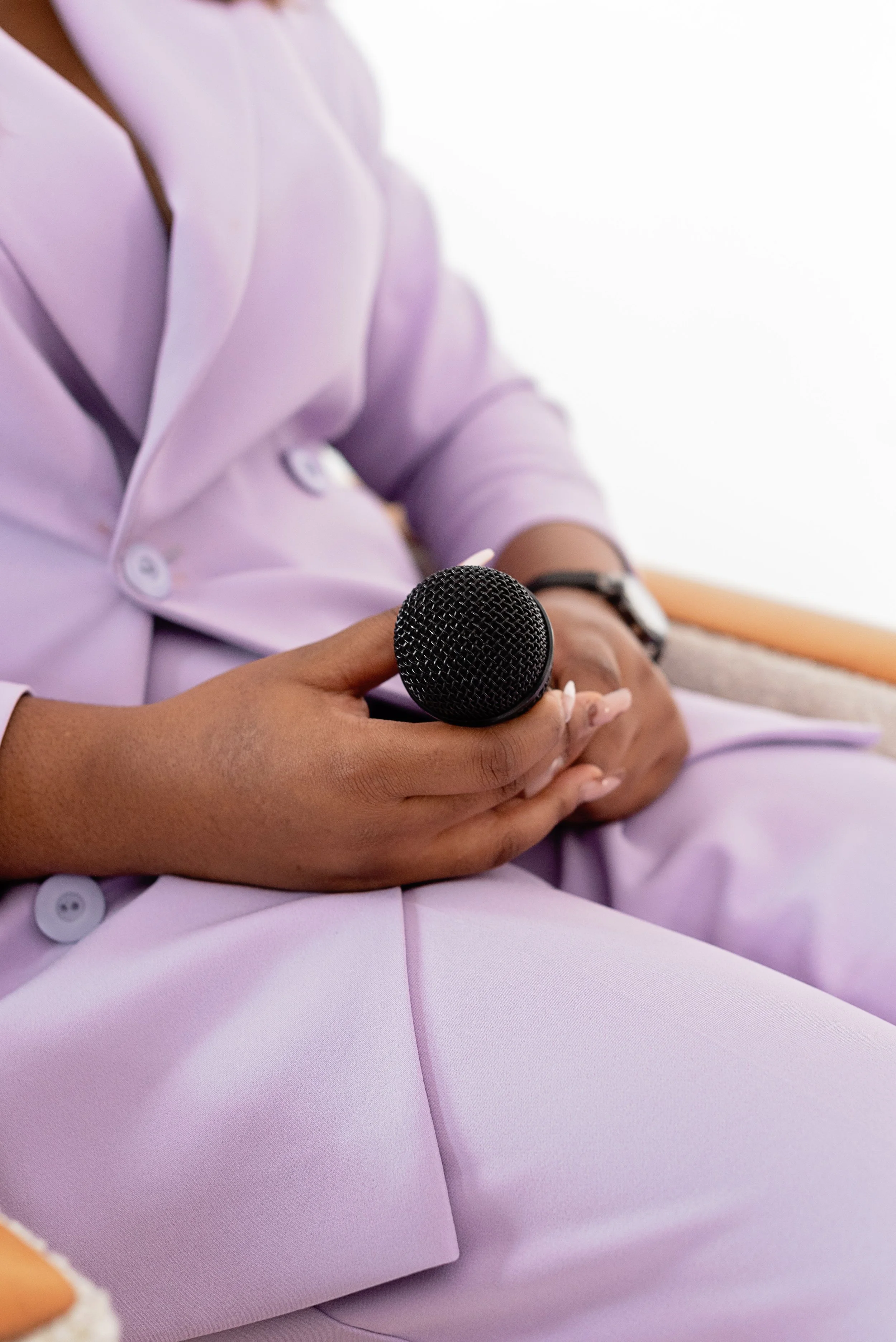 A person wearing a light purple suit holding a black microphone, with a white background.
