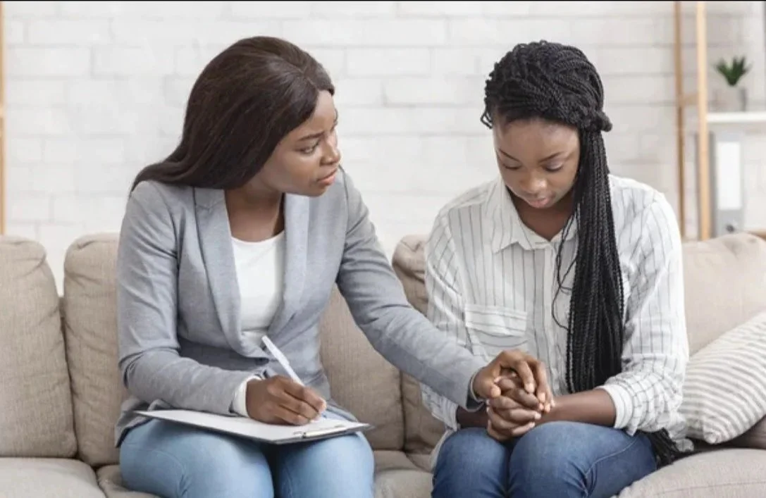 A woman counseling a young girl on a beige couch, with the woman holding the girl's hands and taking notes.