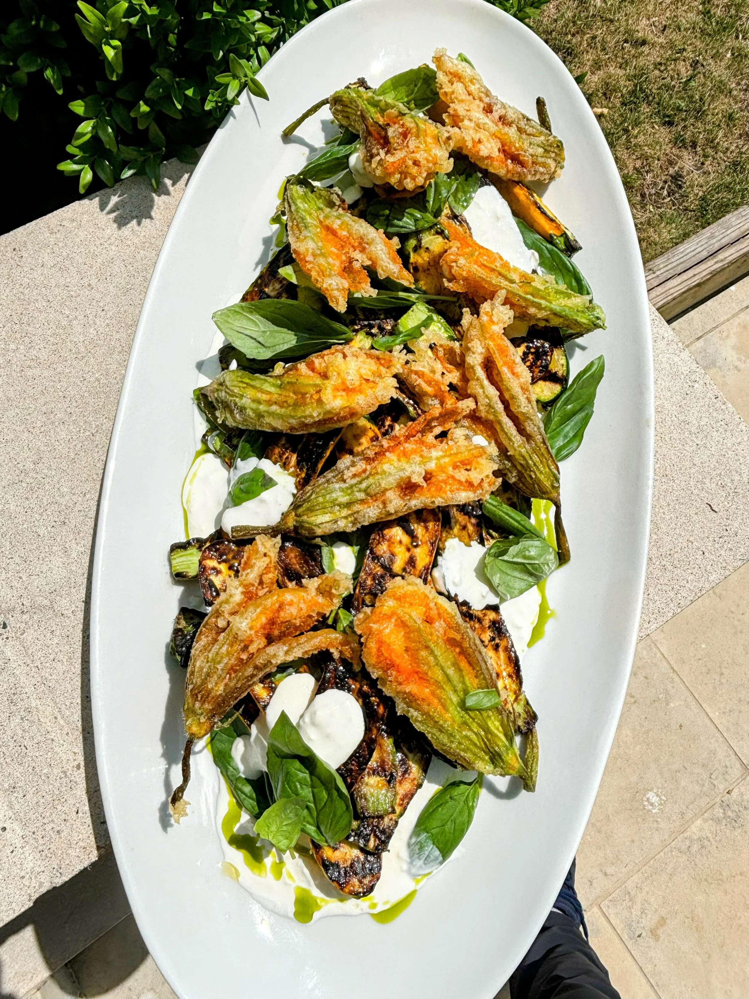Grilled vegetables, including zucchini and eggplant, topped with fried zucchini blossoms, fresh basil leaves, and drizzled with olive oil, served on a white oval platter.