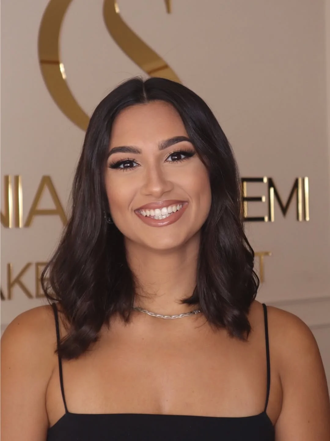 Portrait of a woman with shoulder-length brunette hair, soft glam makeup, and a black outfit, smiling.