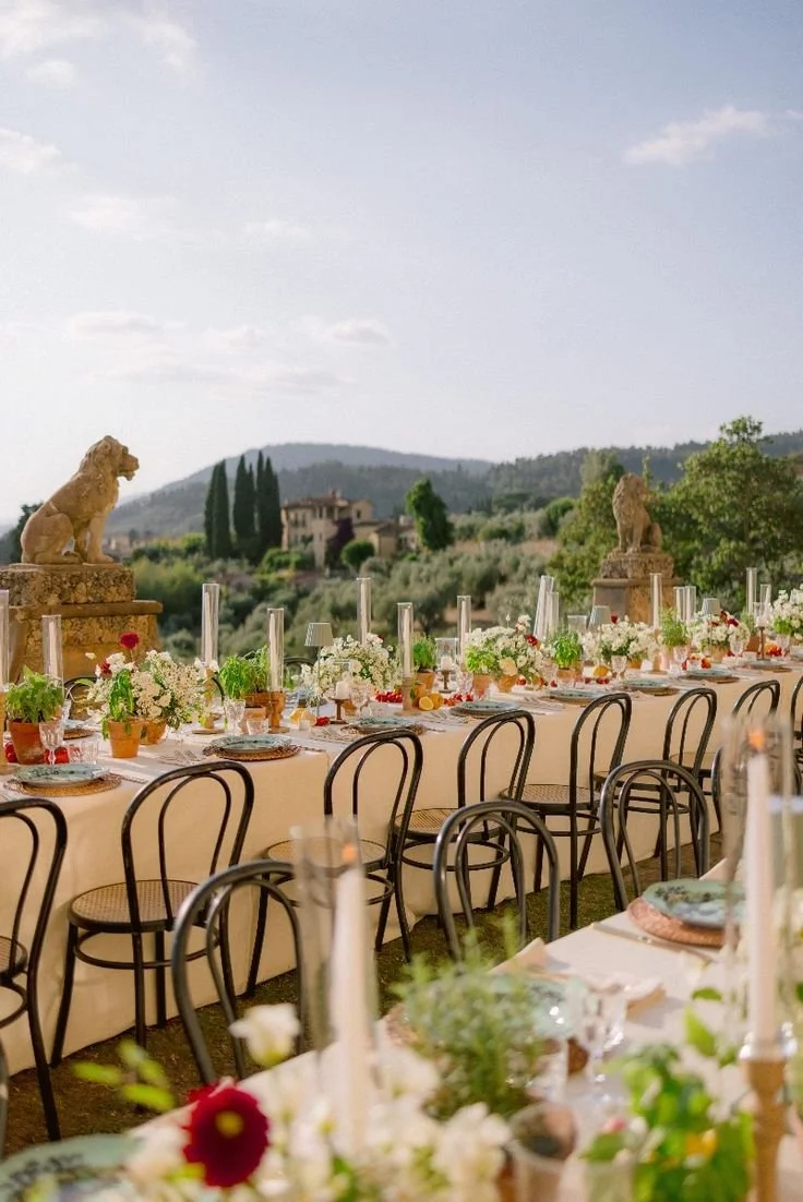 Long outdoor welcome party table in Tuscany with bentwood chairs, white florals, candles, and countryside views.