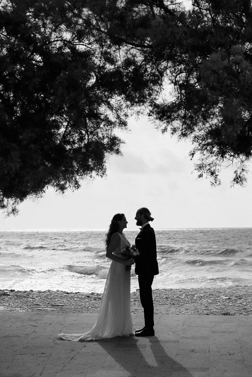 Bride and groom standing beneath trees beside the sea during their Italian wedding, photographed in black and white by Lauren Young.