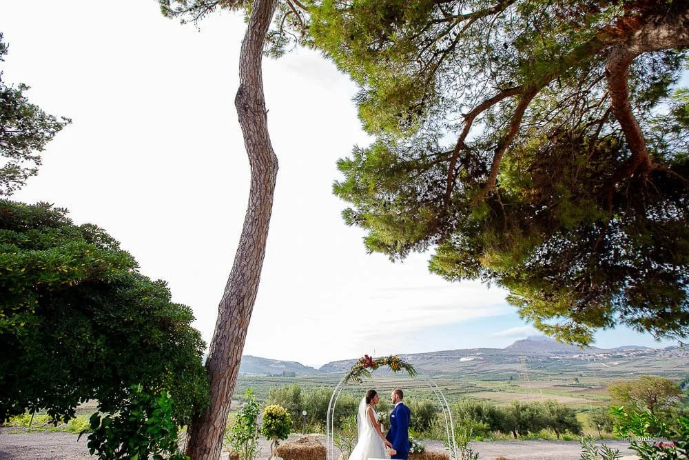 Bride and groom standing under a floral arch during an outdoor countryside wedding ceremony in Sicily with olive trees and mountain views