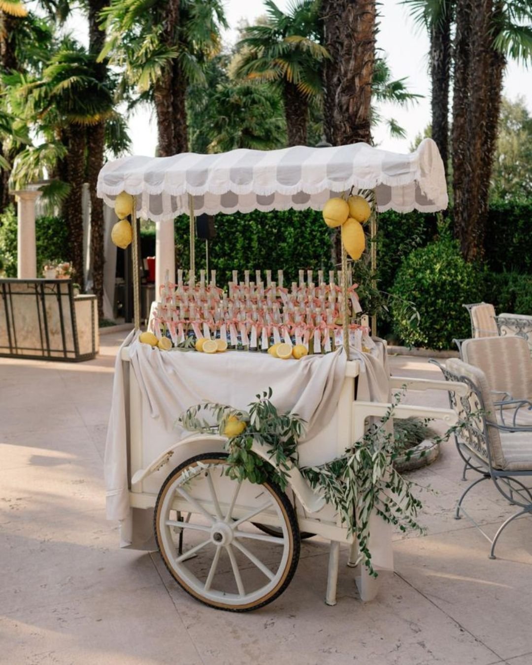 Elegant limoncello cart with fresh lemons and bottled drinks at an outdoor Italian wedding reception surrounded by palm trees.