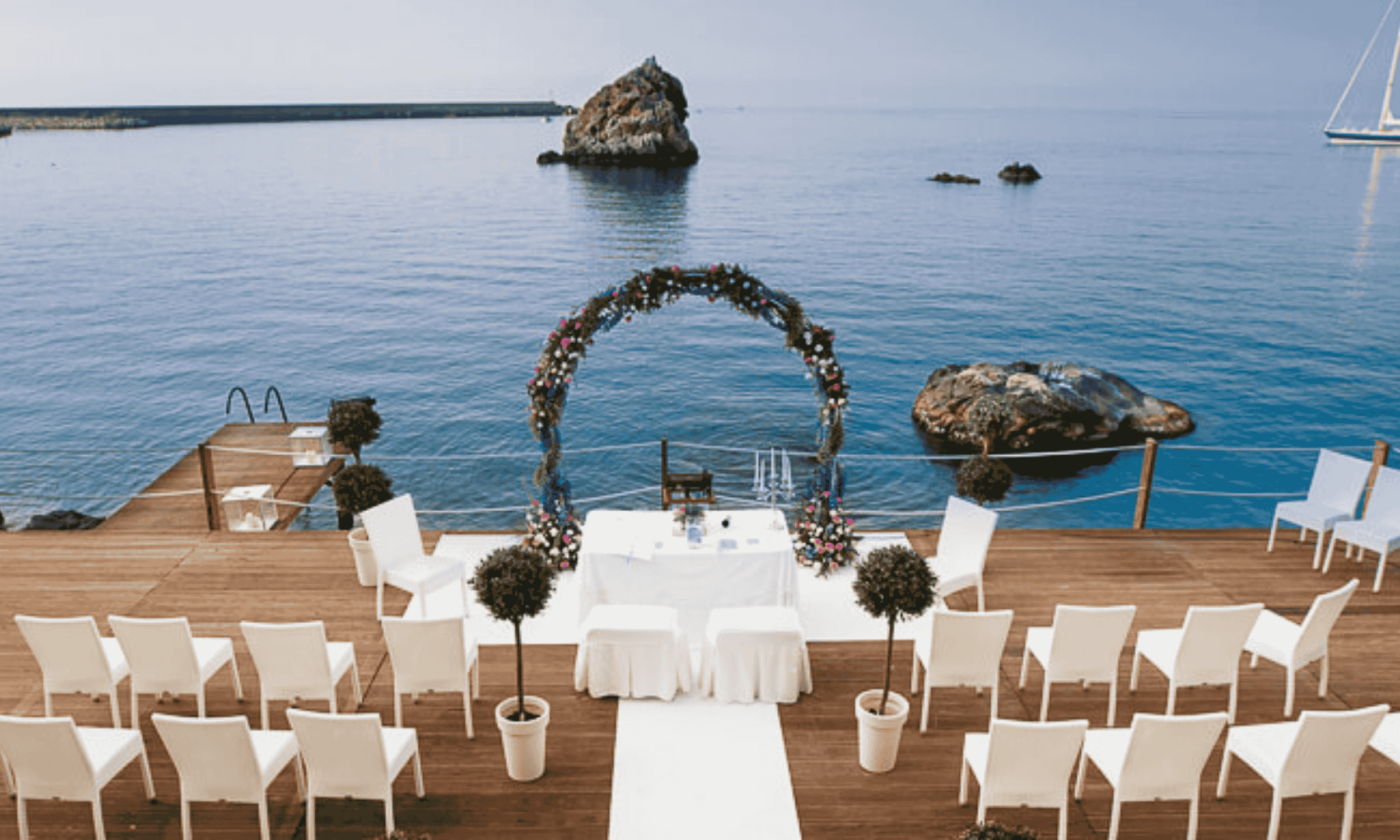 Outdoor seaside wedding ceremony setup in Italy with white chairs and floral arch overlooking the ocean and rocky coastline