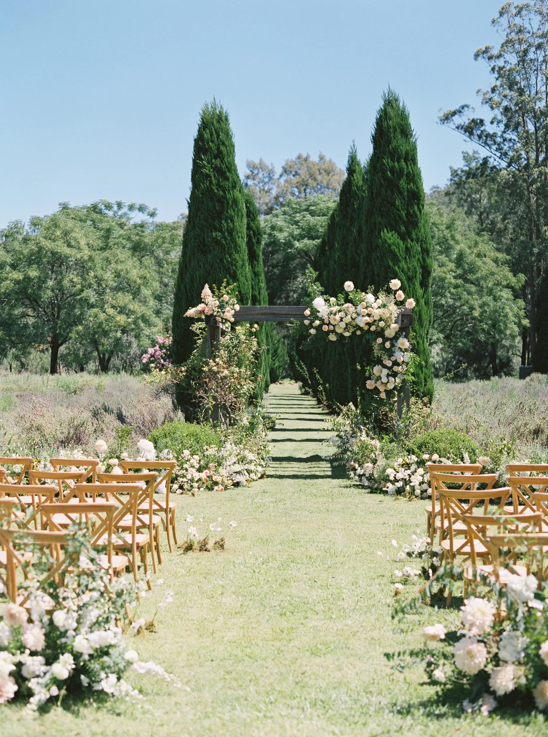 Outdoor garden wedding ceremony set along a symmetrical grass aisle lined with wooden chairs, leading to a floral covered wooden arch framed by tall cypress trees