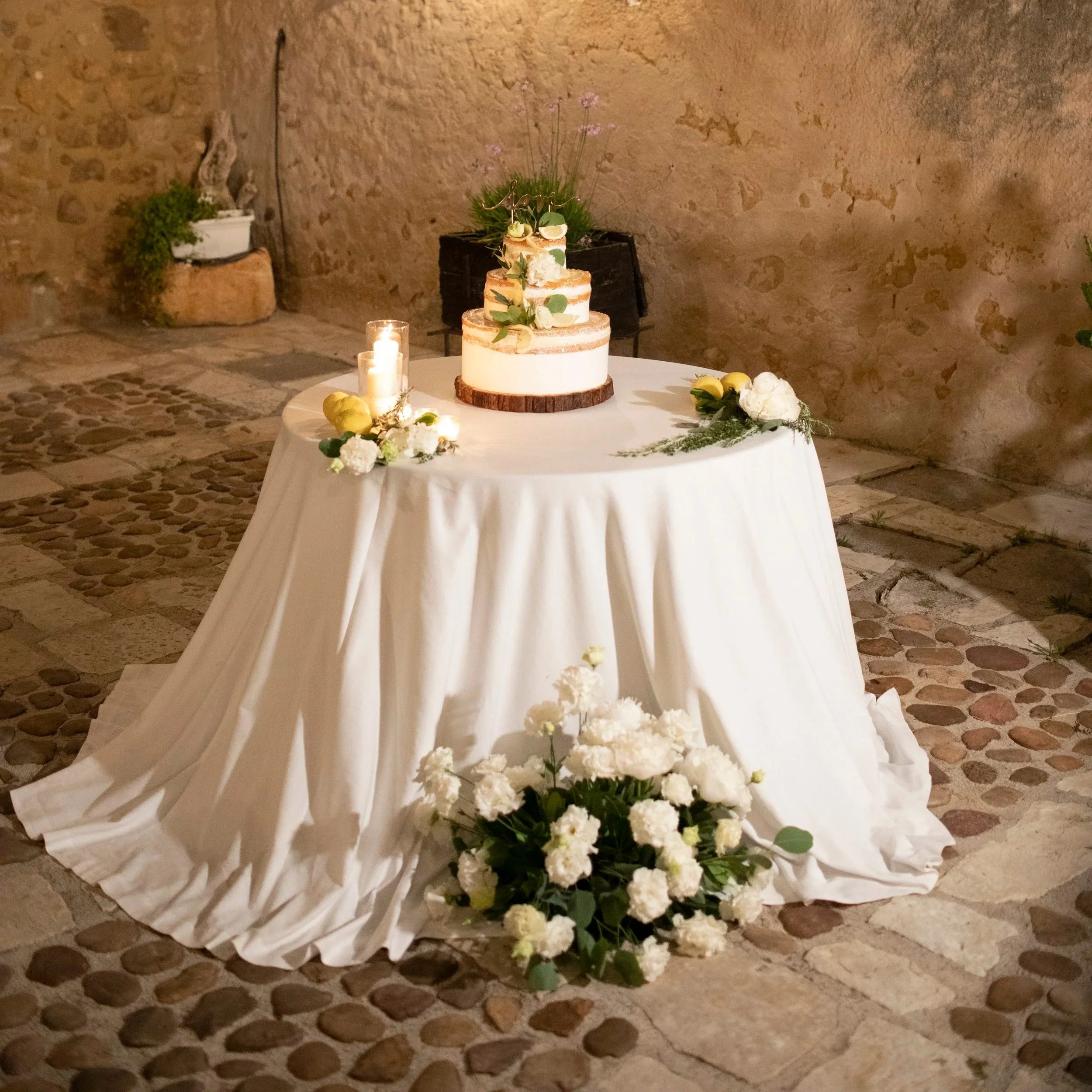 Rustic wedding cake table with candlelight and white floral arrangements in a romantic Sicilian courtyard during an evening wedding reception.