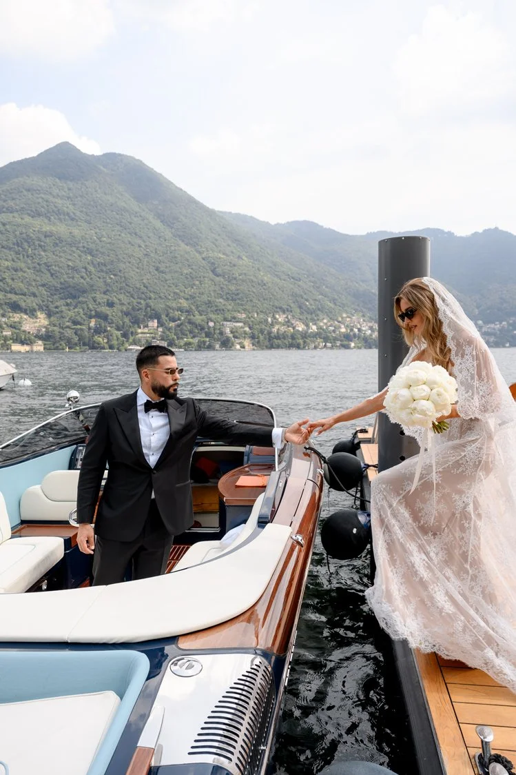 Bride and groom stepping between elegant wooden boats on Lake Como, with green mountains in the background.
