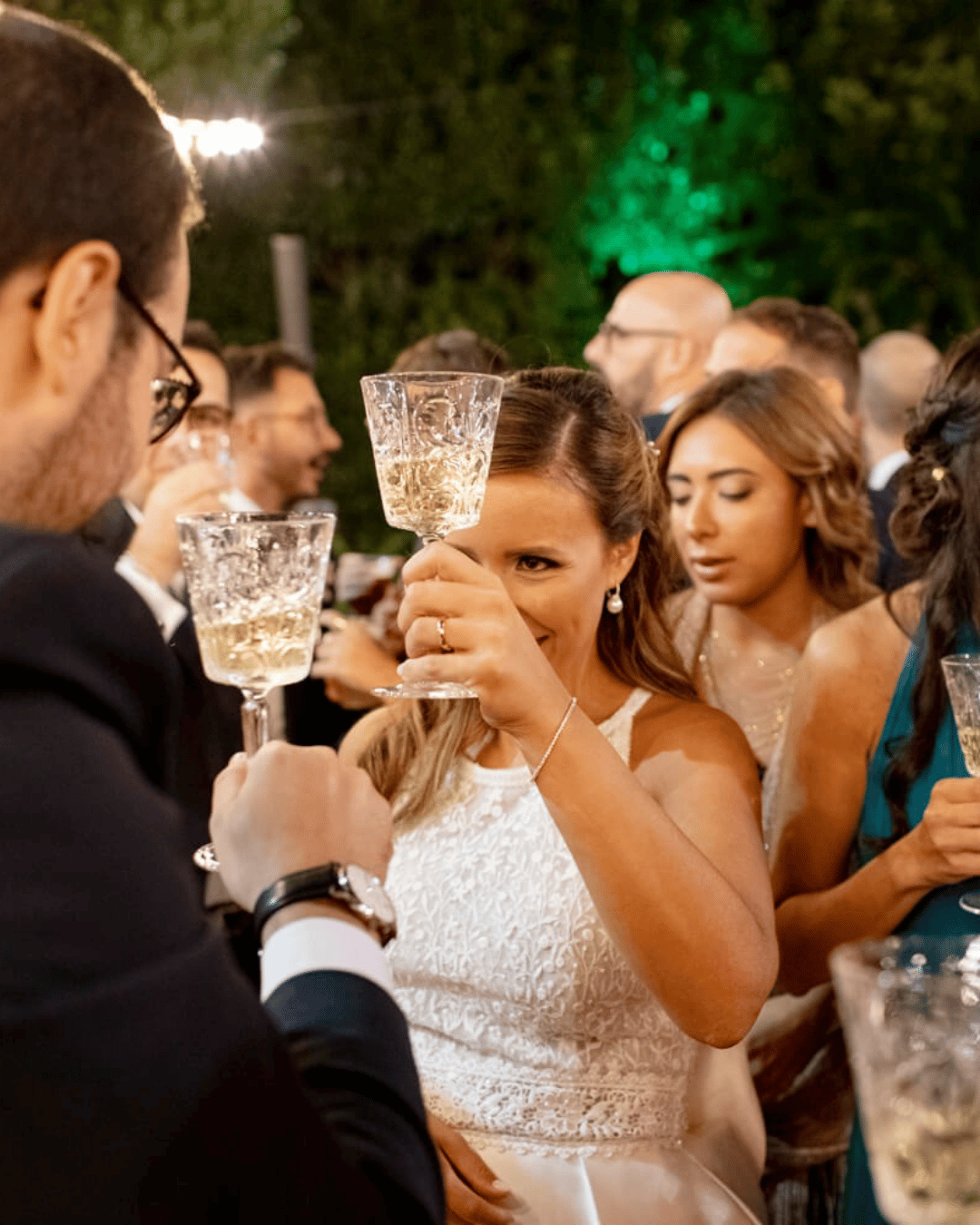 Guests raising glasses and celebrating during an outdoor wedding reception in Sicily, Italy, enjoying drinks beneath festoon lights at an evening wedding celebration.