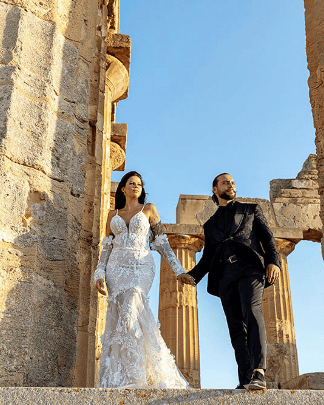 Bride and groom walking through ancient temple ruins in Sicily with golden stone columns and blue sky, romantic destination wedding portrait