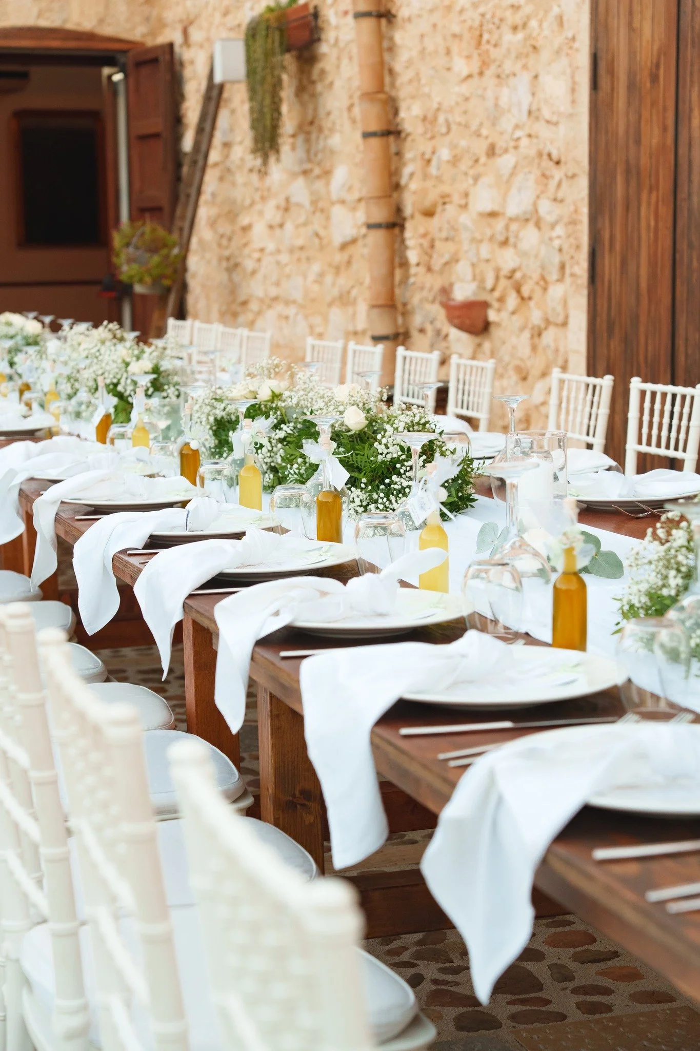 Elegant outdoor wedding banquet table at Baglio Aversa with white chairs, wooden tables, soft floral centrepieces, folded white napkins, and rustic Sicilian courtyard charm.