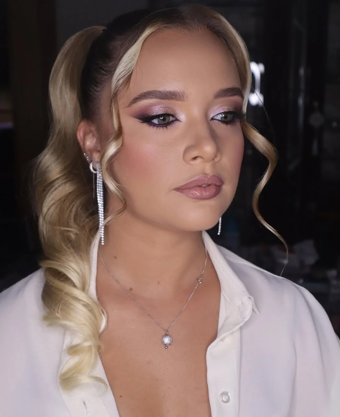 Close-up portrait of a woman with blonde hair in a curled ponytail, soft glam bridal makeup, and elegant jewellery.