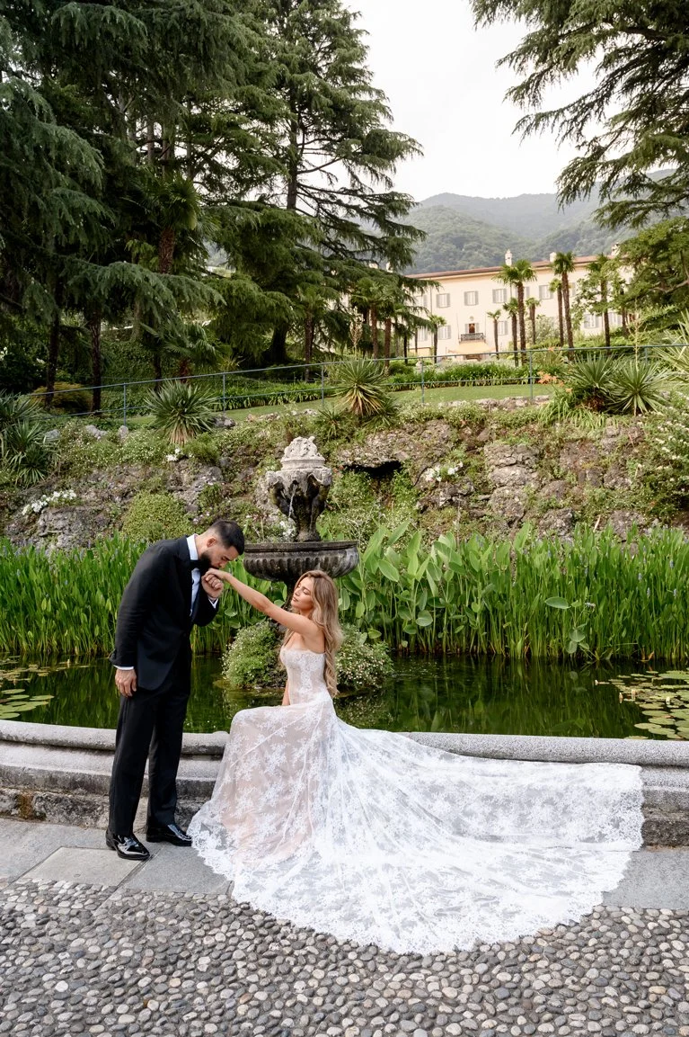 Groom kissing the bride’s hand beside a garden pond at a Lake Como wedding venue, with her lace train spread across the stone path.