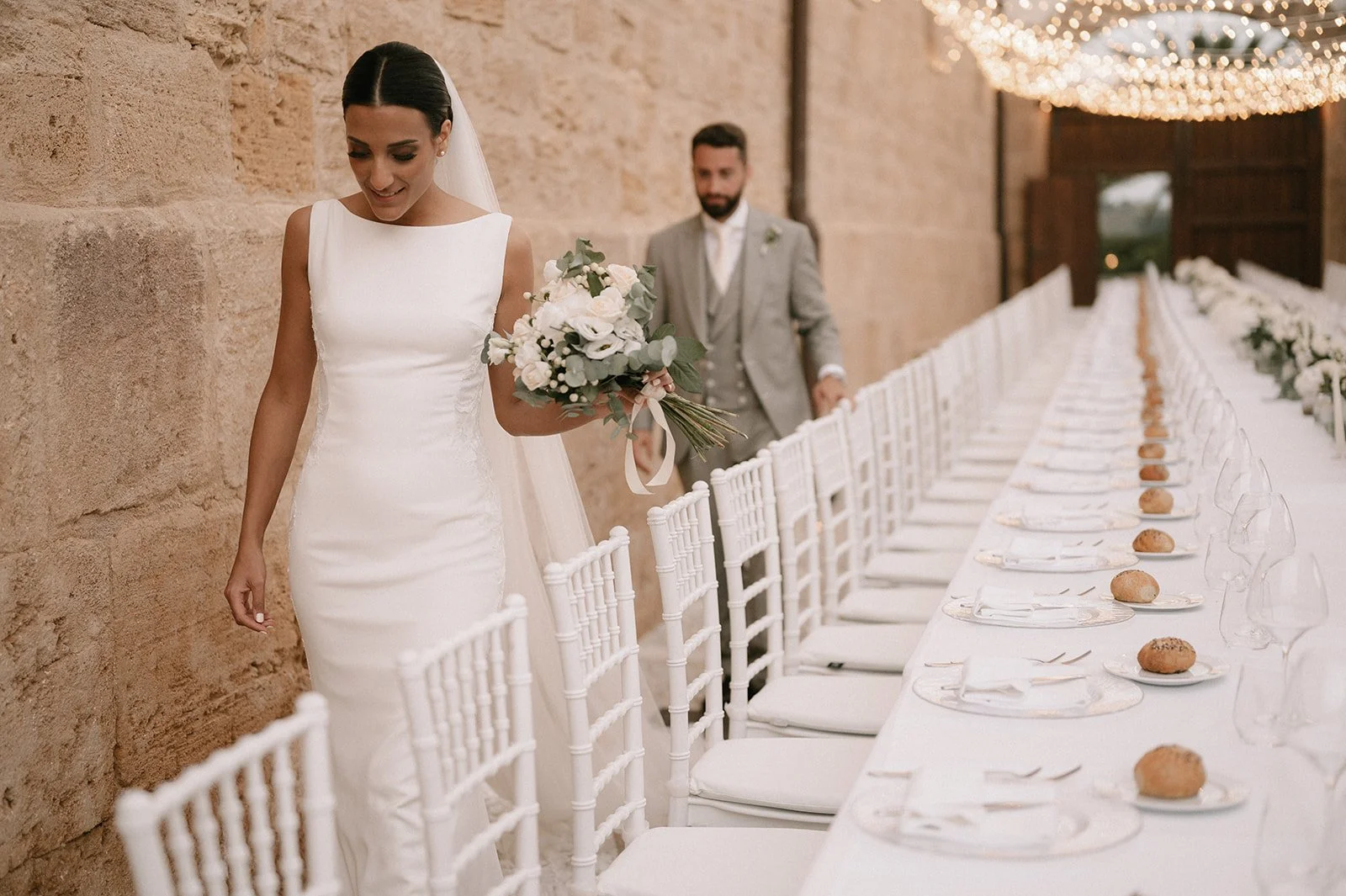 Bride holding a bouquet while walking beside elegant reception tables styled with white florals and candles.