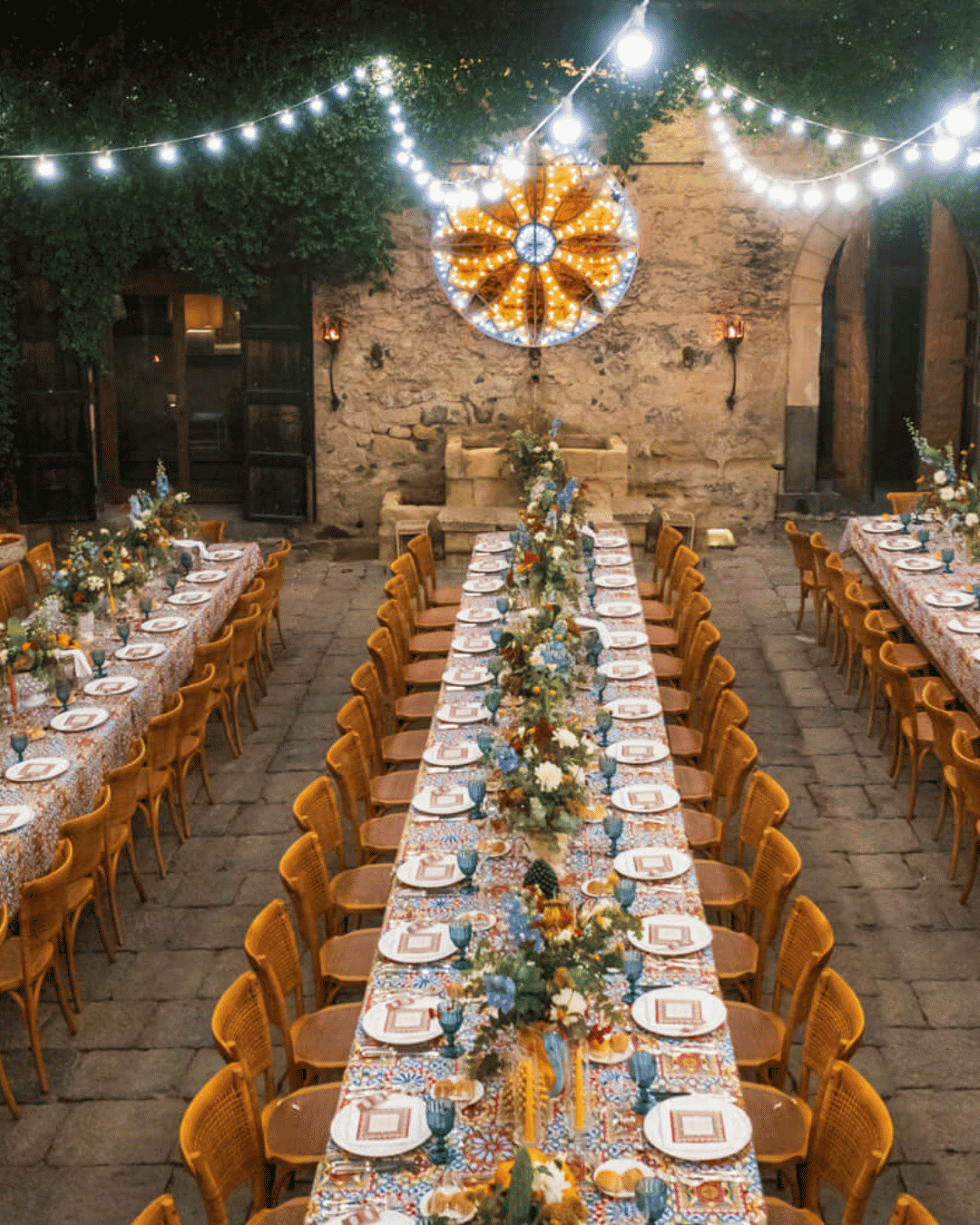 Long banquet tables set for an outdoor wedding reception in a historic Sicilian courtyard, decorated with colourful florals and festoon lights for an Italian destination wedding.