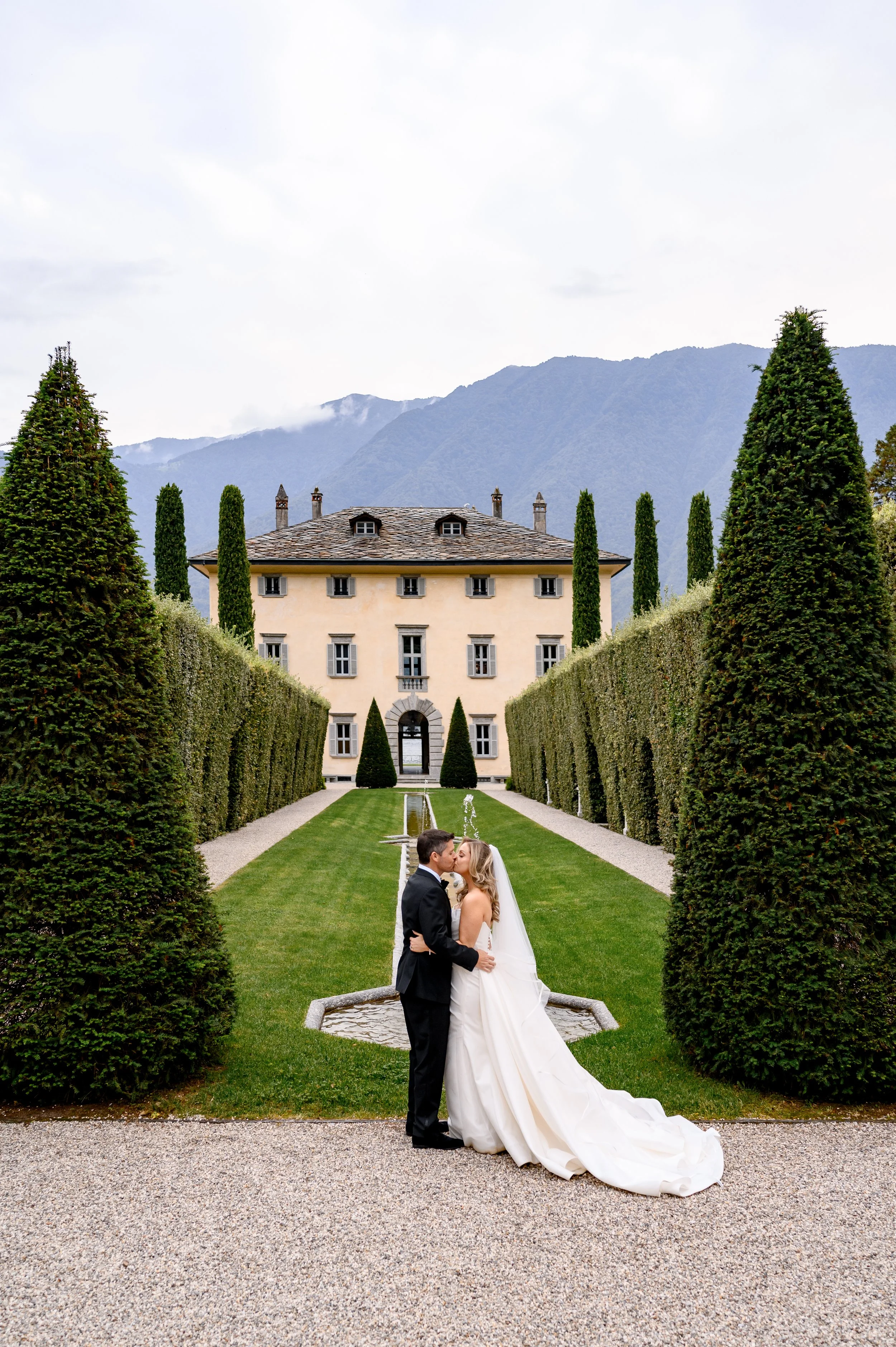 Bride and groom sharing a kiss in the formal gardens of a historic Italian villa surrounded by cypress trees and mountain views.