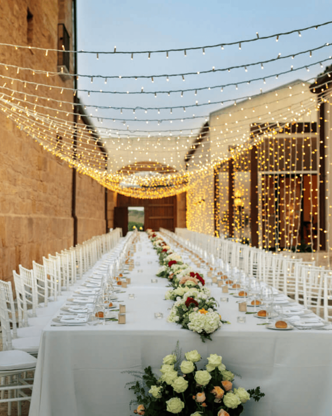 Long elegant wedding reception table in an Italian courtyard with white chairs, floral arrangements and overhead fairy lights at dusk