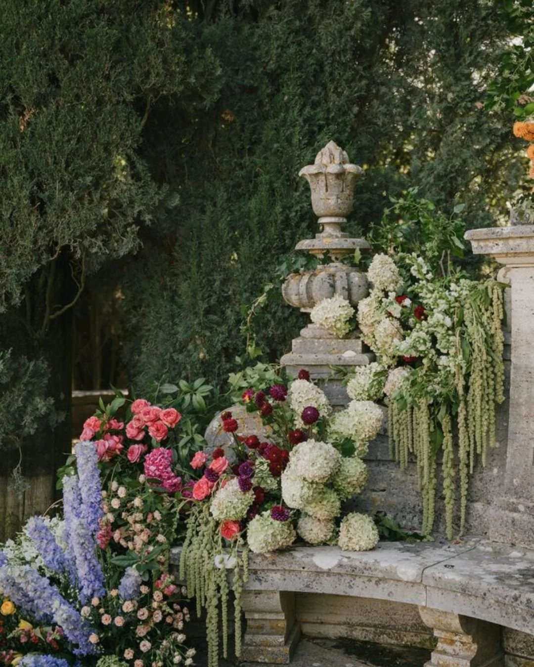 Lush garden wedding floral installation on a stone balustrade with white hydrangeas, pink roses, burgundy blooms, and trailing greenery against tall trees.