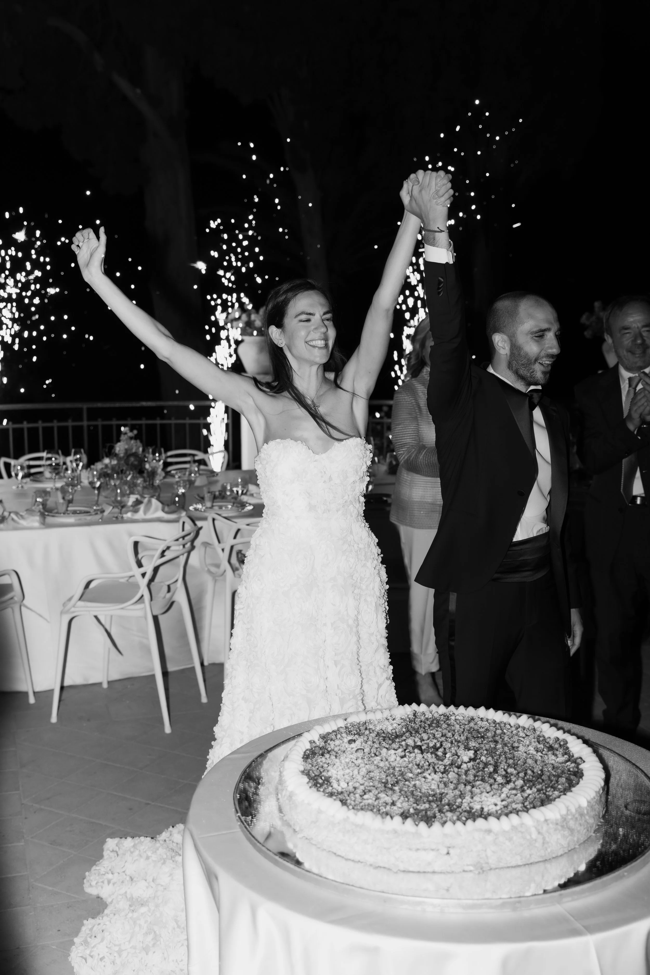 Newlyweds celebrating beside their wedding cake with sparklers during an Italian reception, photographed in black and white by Lauren Young.