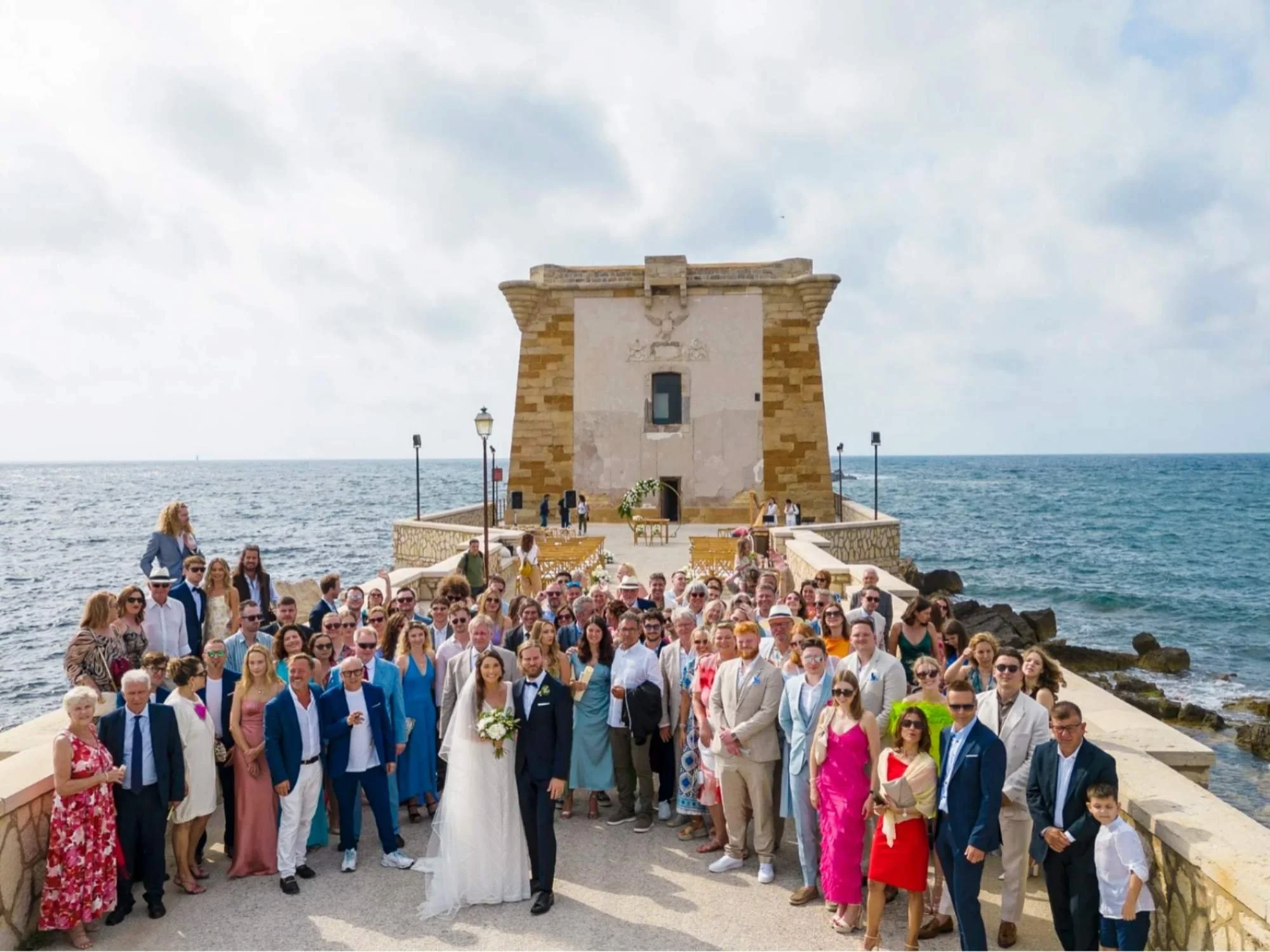 A joyful group photo of family and friends in front of Torre di Ligny, celebrating love by the sea.