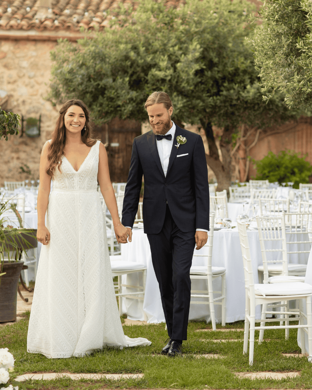 Couple walking through outdoor wedding reception setup in Italy with white tables, rustic venue, and olive trees