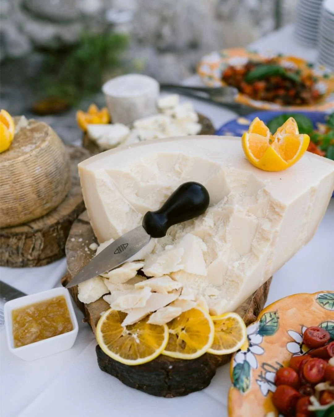 Large wheel of aged Italian cheese displayed on a rustic wooden board with lemon slices, honey, and cherry tomatoes at an outdoor wedding aperitivo.”