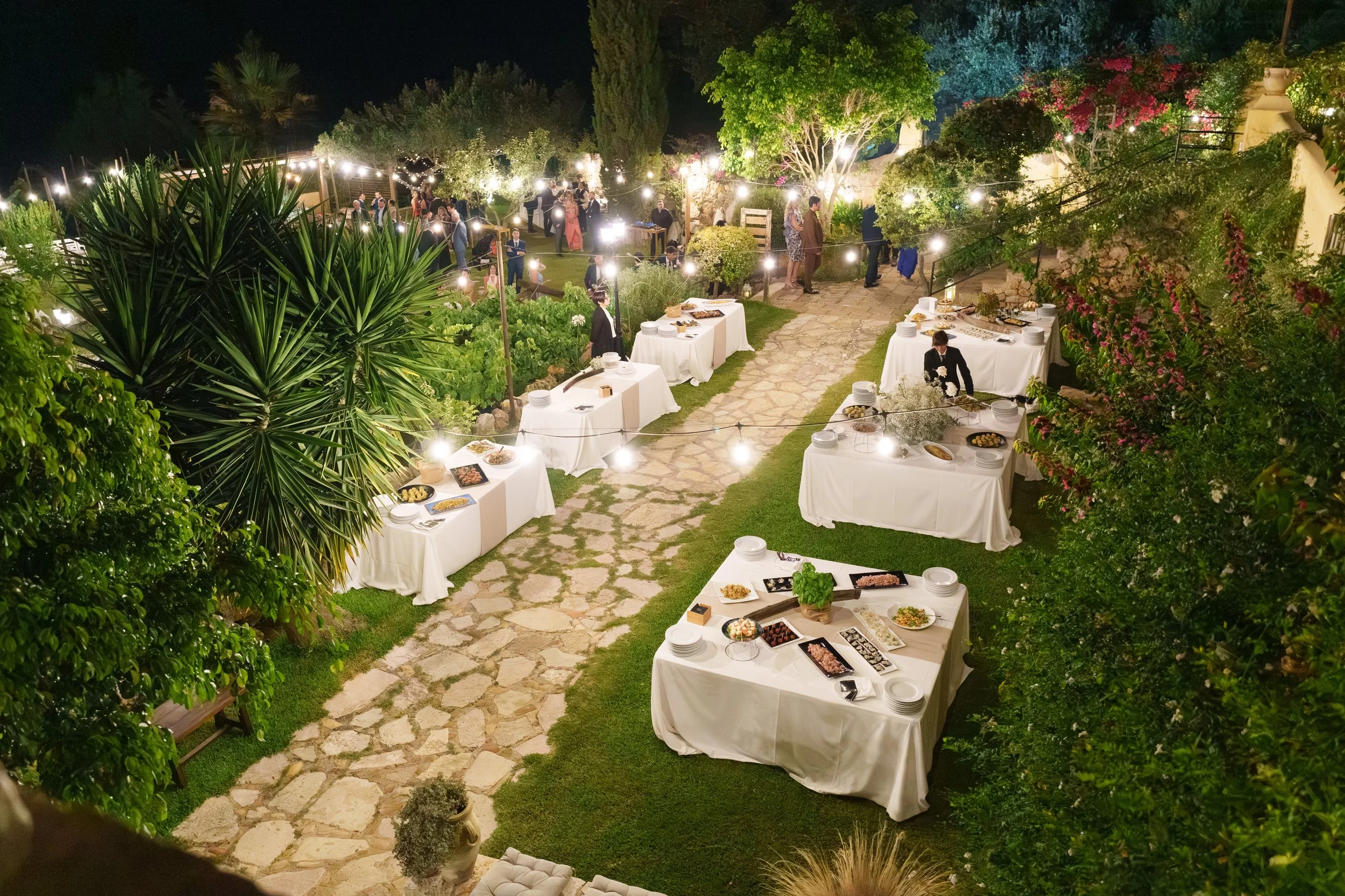 An aerial night view of an outdoor wedding reception in an Italian garden, with guests dining at long white-clothed tables under warm string lights surrounded by lush tropical plants.