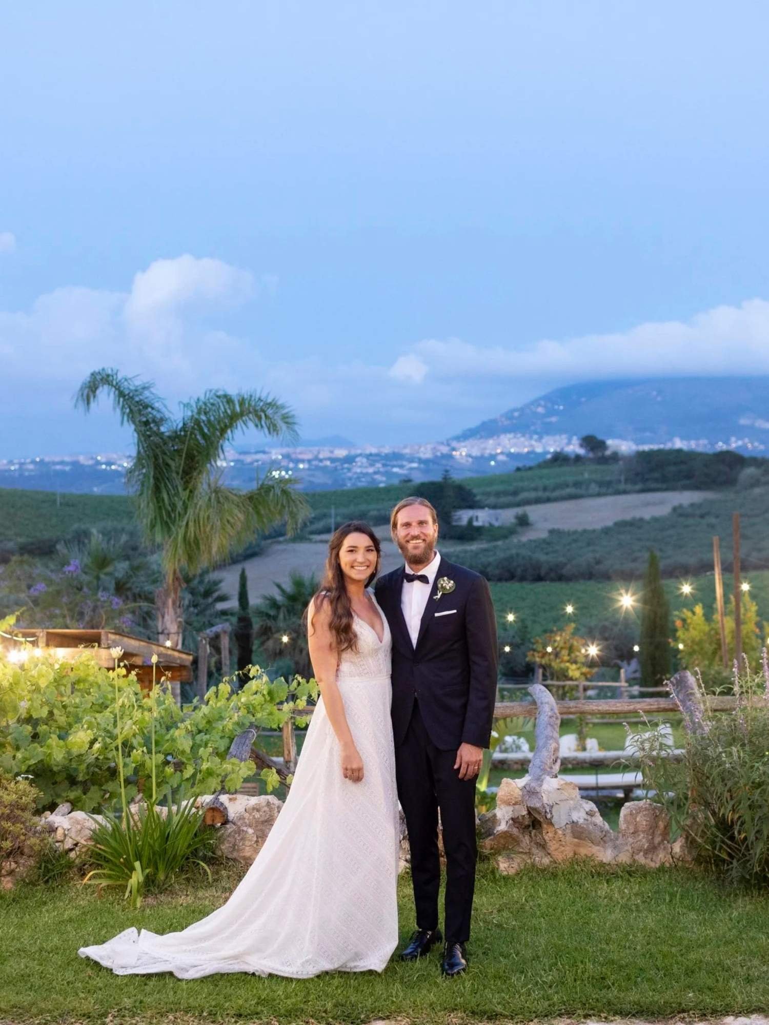 Newlyweds smiling at sunset in the vineyards of Baglio Aversa, surrounded by twinkling string lights and sweeping Sicilian countryside views.