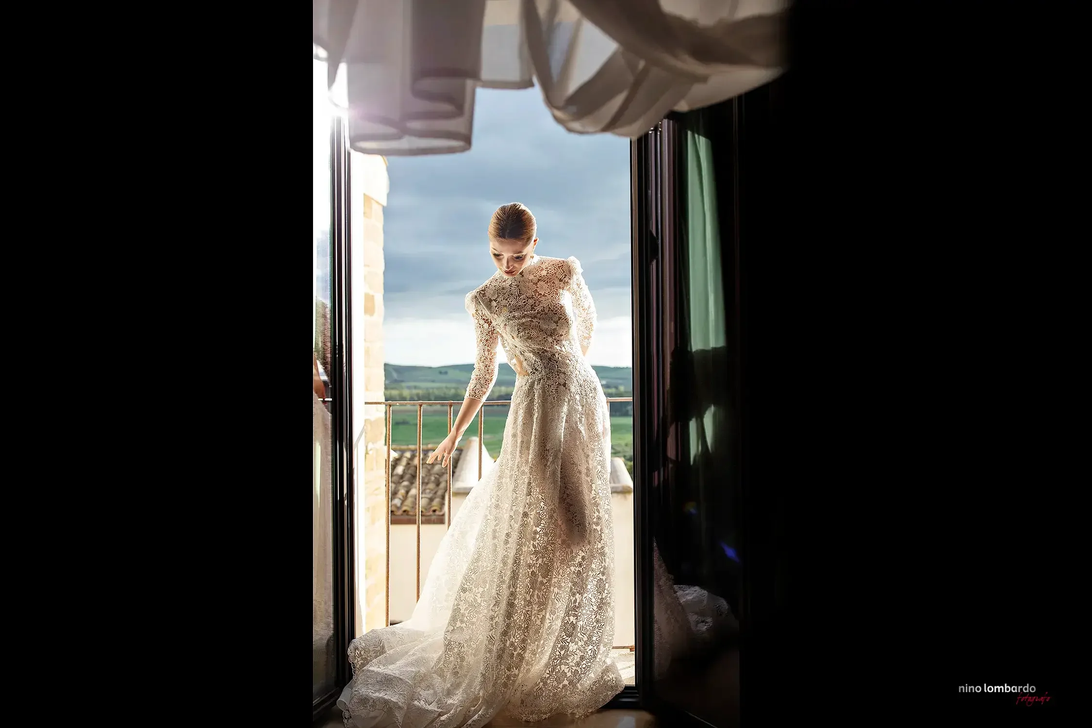 Bride in lace wedding dress standing in a doorway with soft natural light and countryside views in Sicily