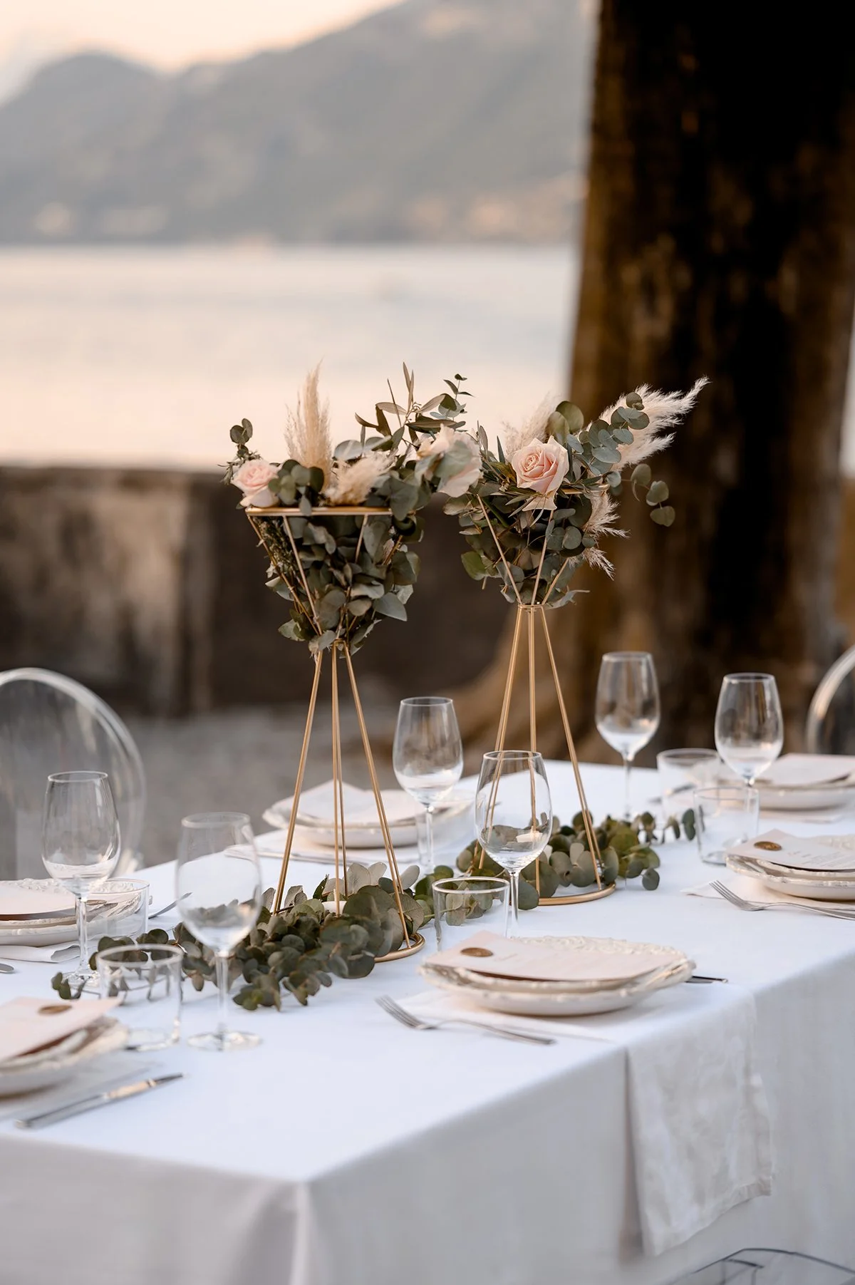Elegant wedding table styling on Lake Como featuring gold stands, soft blush roses, eucalyptus greenery, and lake views in the background