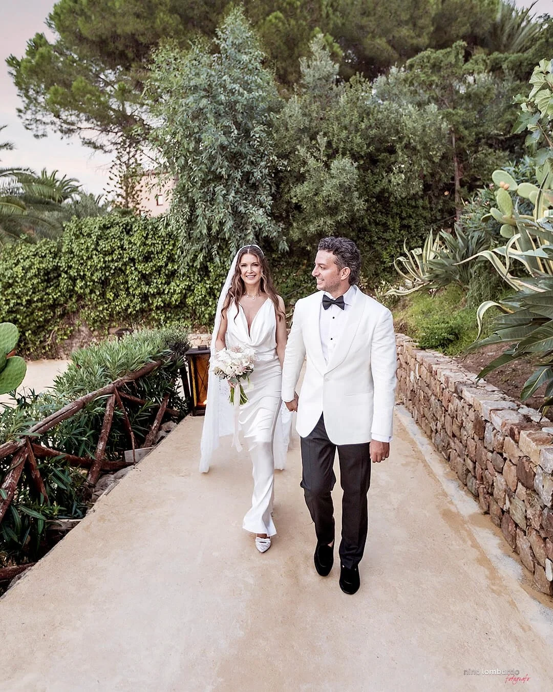 Bride and groom walking hand in hand along a garden path in Sicily surrounded by greenery and rustic stone walls