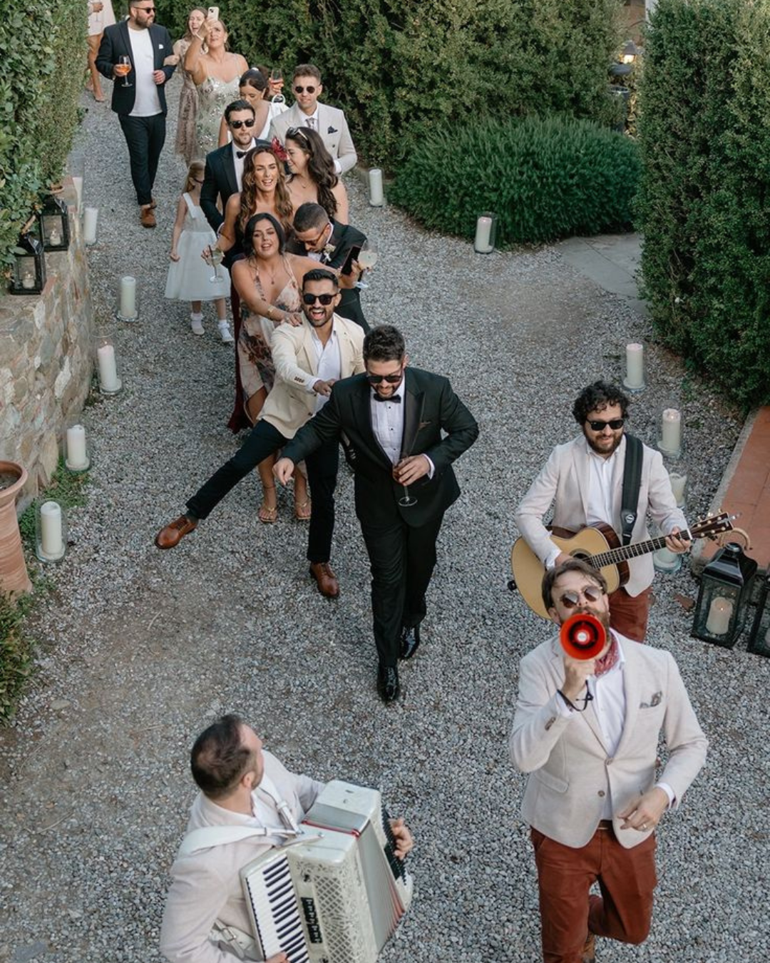 Guests dancing in a joyful conga line behind live musicians at an Italian wedding, with accordion, guitar, and megaphone leading the way through a garden path.