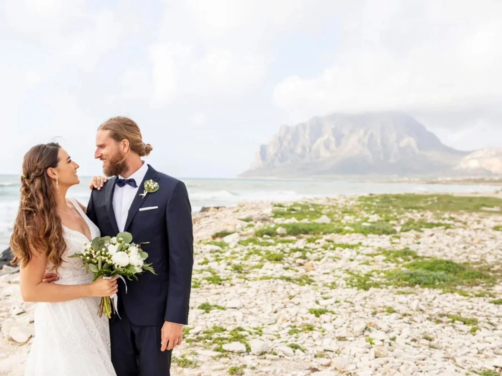 Bride and groom sharing a loving gaze on the rocky coastline of Trapani, with the majestic Sicilian mountains rising in the background.