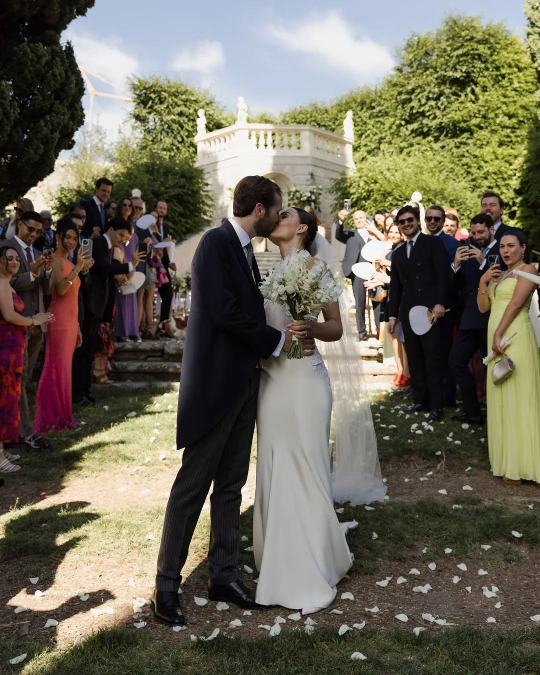 Bride and groom kissing after their outdoor wedding ceremony in Italy, surrounded by cheering guests in a villa garden.