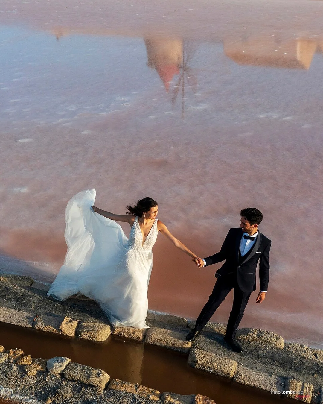 Bride and groom holding hands walking along stone path beside a pink salt lake in Sicily with soft sunset light