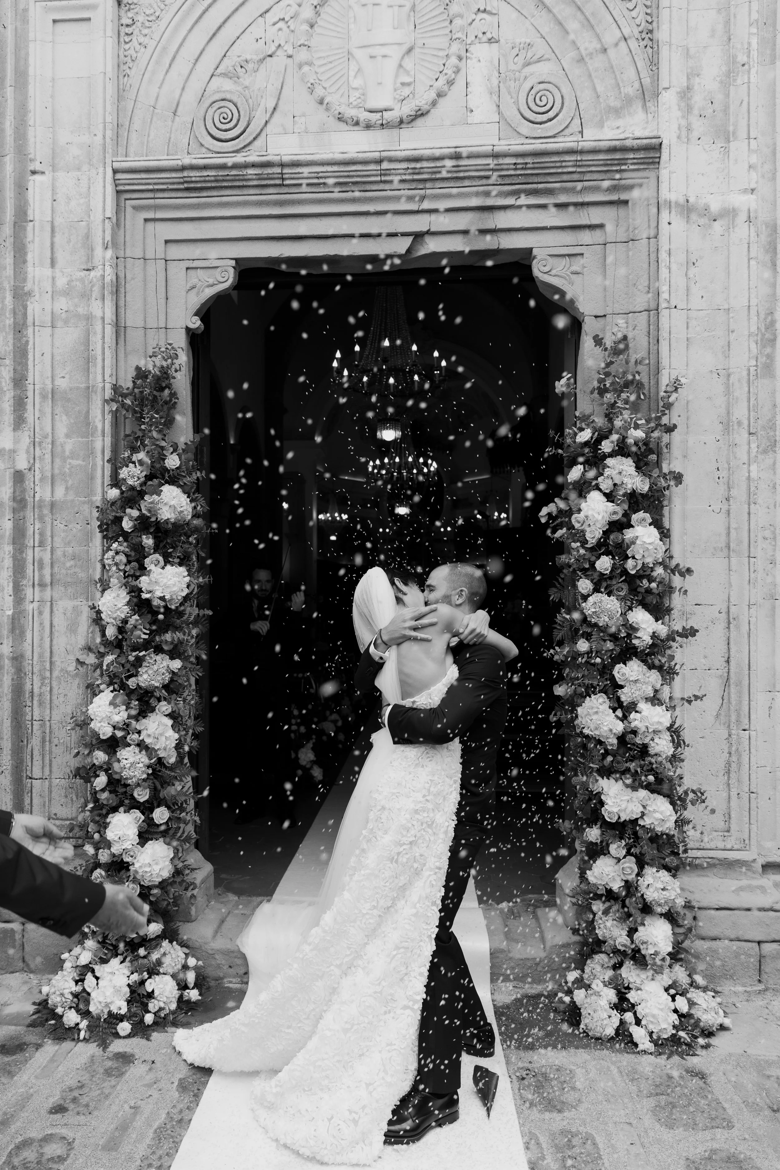 Newlyweds embracing under falling confetti outside a flower filled Italian church, photographed in black and white by Lauren Young.
