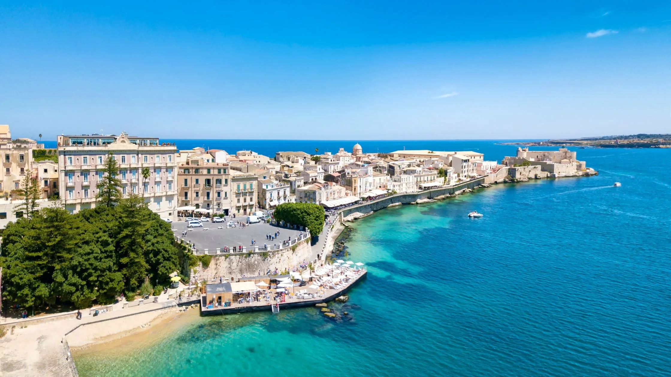 Aerial view of Ortigia Island in Syracuse, Sicily, with historic buildings along the coastline and turquoise Mediterranean waters.